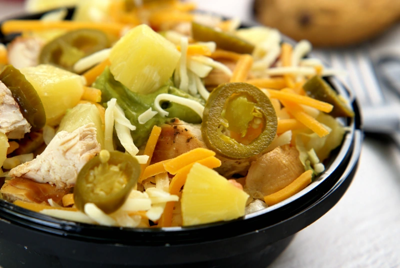Close-up of a chicken rice bowl with jalapeños, pineapple, shredded cheese, and guacamole in a black container